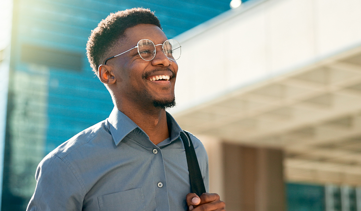 A smiling young man in glasses and a gray shirt walks outside a modern building
