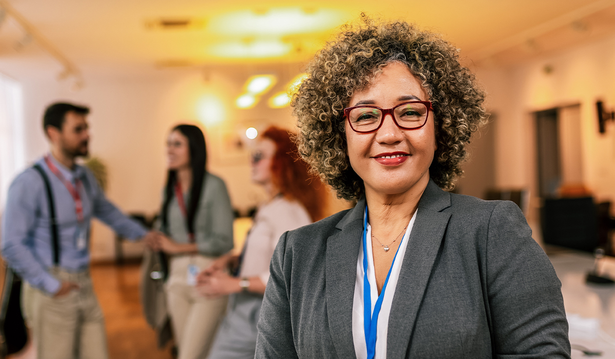 A woman in a gray blazer and red glasses smiles in the foreground while colleagues network in the background