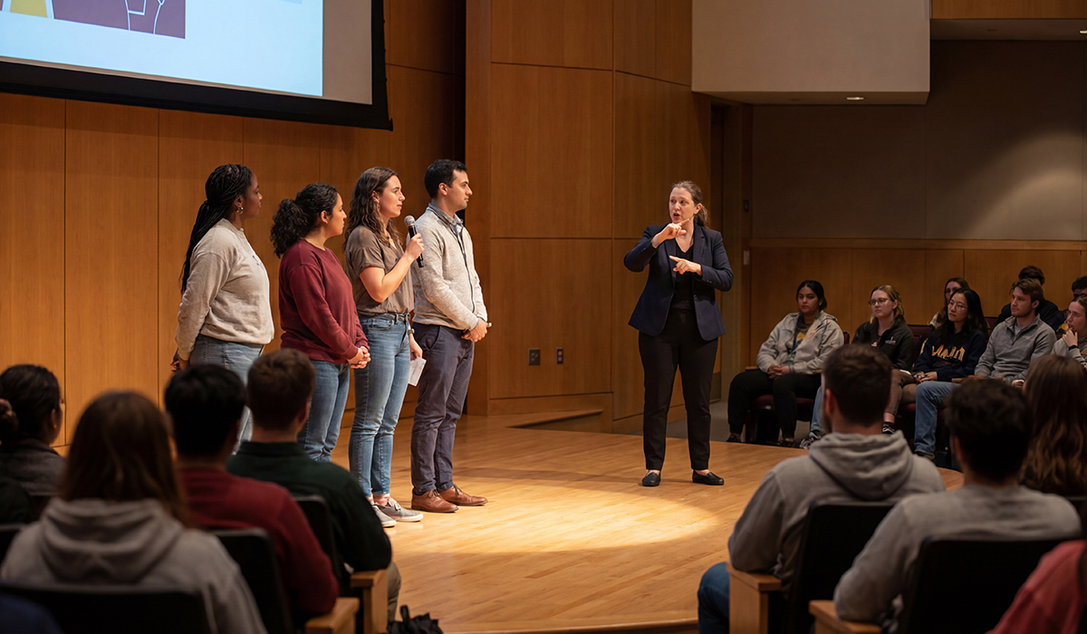 Four speakers present on stage in a wood-paneled auditorium while a sign language interpreter signs next to them for an audience.