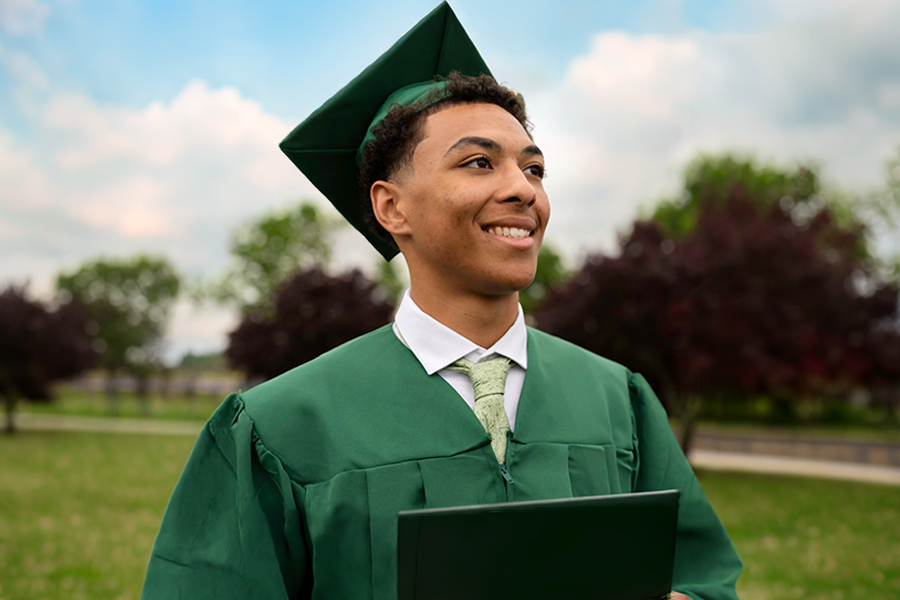 A smiling graduate in a green cap and gown holds his diploma and gazes into the distance on a sunny day.