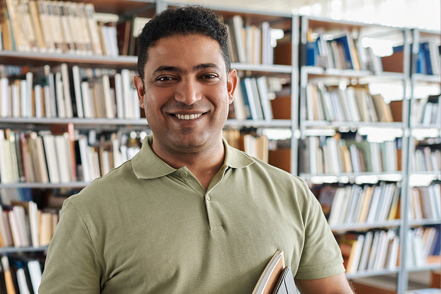A smiling adult man standing in a library holding books, with shelves of books visible behind him.