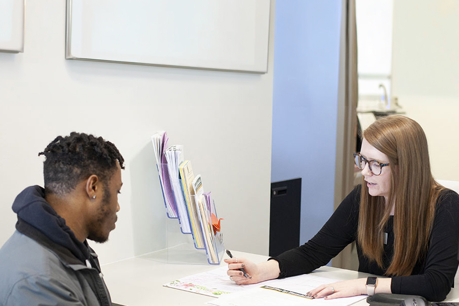 Alt Text: A woman with long brown hair and glasses sits at a desk, reviewing a document with a young man. She is holding a pen and speaking while gesturing toward the paper. A clear brochure rack filled with pamphlets sits on the desk between them.