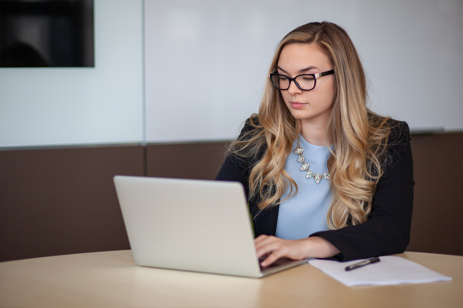 A woman with long blonde wavy hair and black-rimmed glasses sits at a light-colored wooden desk, focused on her laptop. She wears a blue blouse, a black blazer, and a statement necklace.