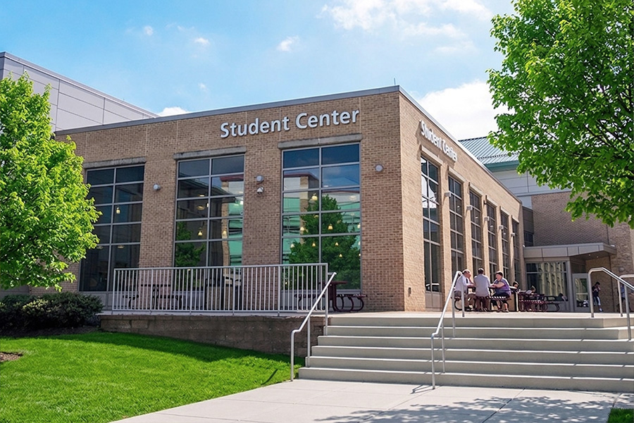 The exterior of the FCC Student Center on a sunny day, with students seated at outdoor tables and green trees framing the brick building.