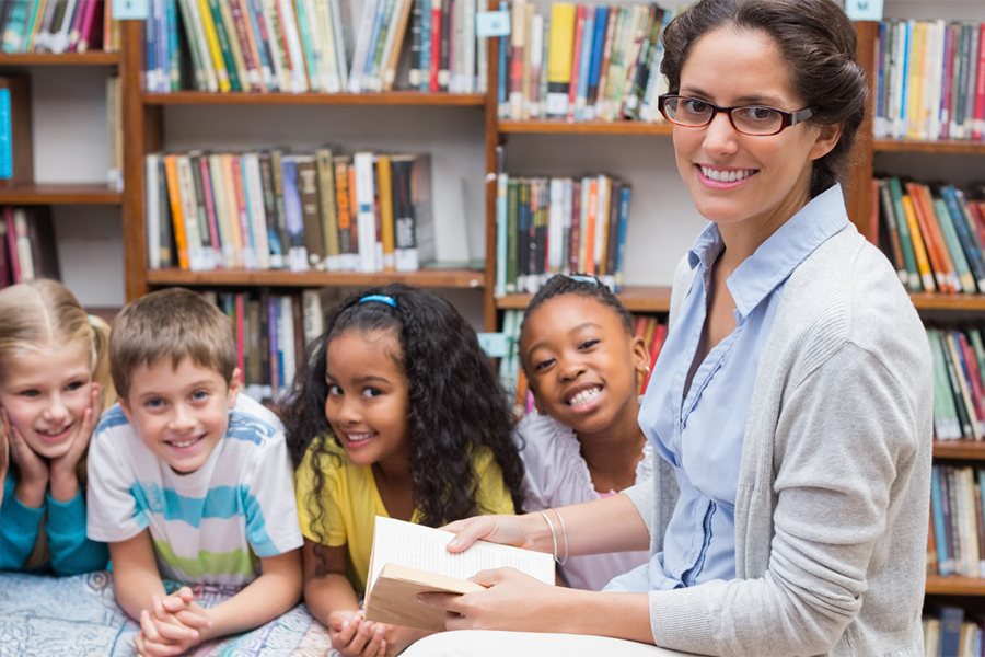 A smiling teacher wearing glasses holds an open book while four young children gather around her attentively in a colorful library setting with bookshelves in the background.