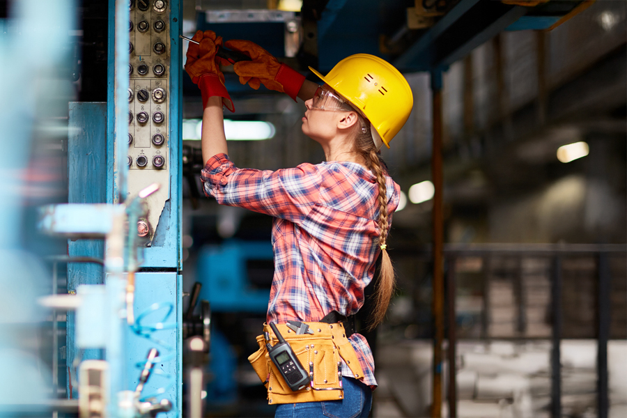 A female electrician in a yellow hard hat, safety glasses, work gloves, and a tool belt works on an electrical control panel in an industrial facility.