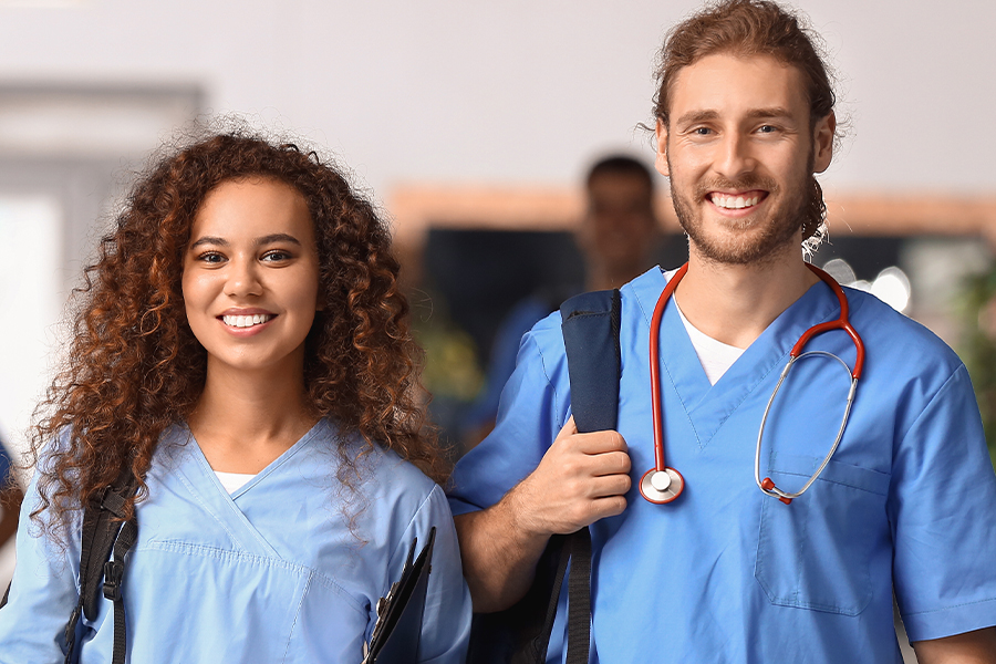 Two smiling healthcare students in blue scrubs carry backpacks in a hallway, with one wearing a red stethoscope around his neck.