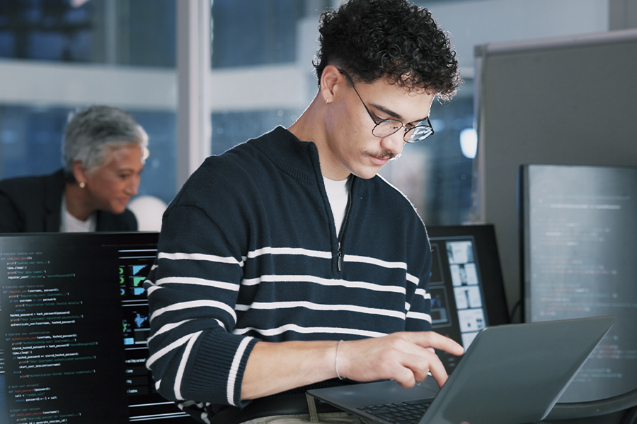 A young man with glasses and curly hair types on a laptop surrounded by monitors displaying lines of code in a dimly lit tech office, with a colleague visible in the background.