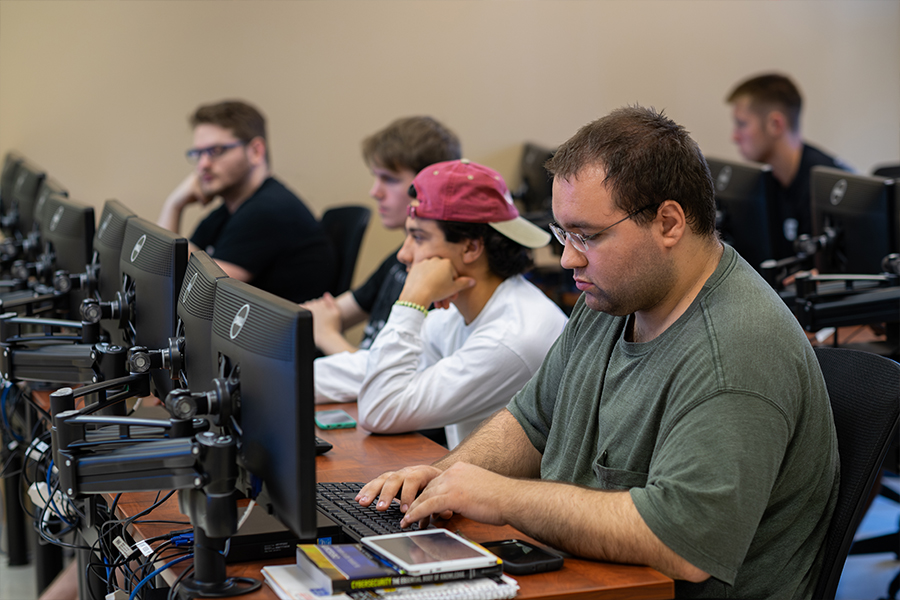 A group of students sits at a row of computer workstations in a classroom. The student in the foreground focuses on a monitor while typing on a keyboard.