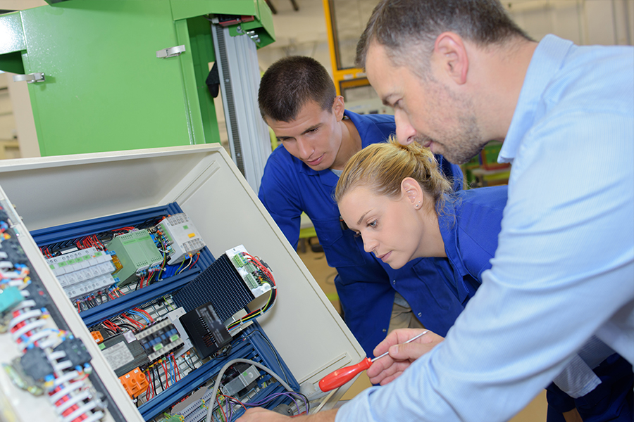 An instructor and two apprentices in blue uniforms lean in closely to examine the wiring inside an open electrical panel during a hands-on training session.