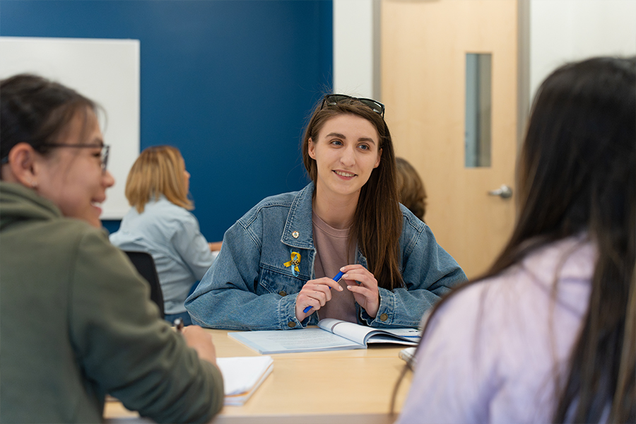 A smiling student in a denim jacket sits at a table with classmates, holding a pen over an open notebook in a bright classroom setting.