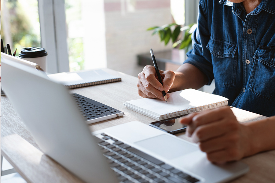 A person in a denim shirt takes notes with a pencil in a notebook while sitting at a desk with a laptop.