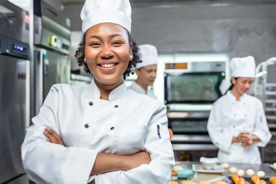 A smiling female pastry chef in a white uniform and toque standing in a professional bakery kitchen.