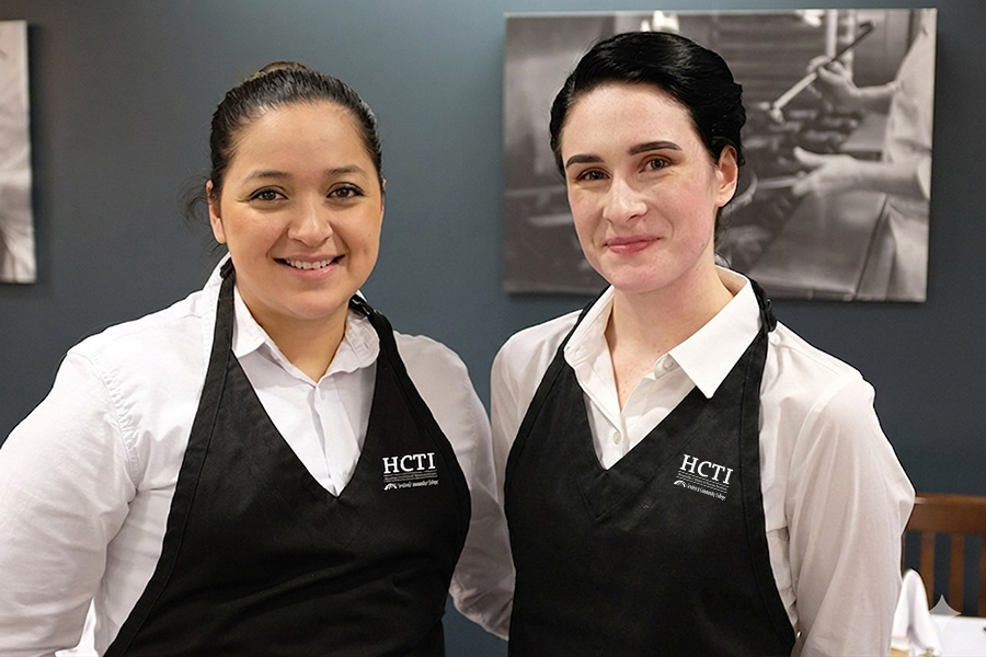 Two smiling FCC HCTI students in white shirts and black HCTI-branded aprons pose together in a dining room setting, with black and white culinary photography visible on the wall behind them.