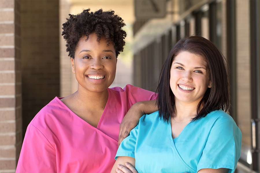 Two healthcare workers in scrubs smile together outdoors in front of a brick building, with one resting their arm on the other's shoulder.