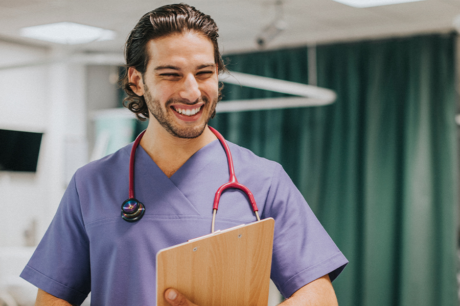 A smiling healthcare worker in purple scrubs and a stethoscope holds a clipboard in a hospital room with green privacy curtains visible behind them.
