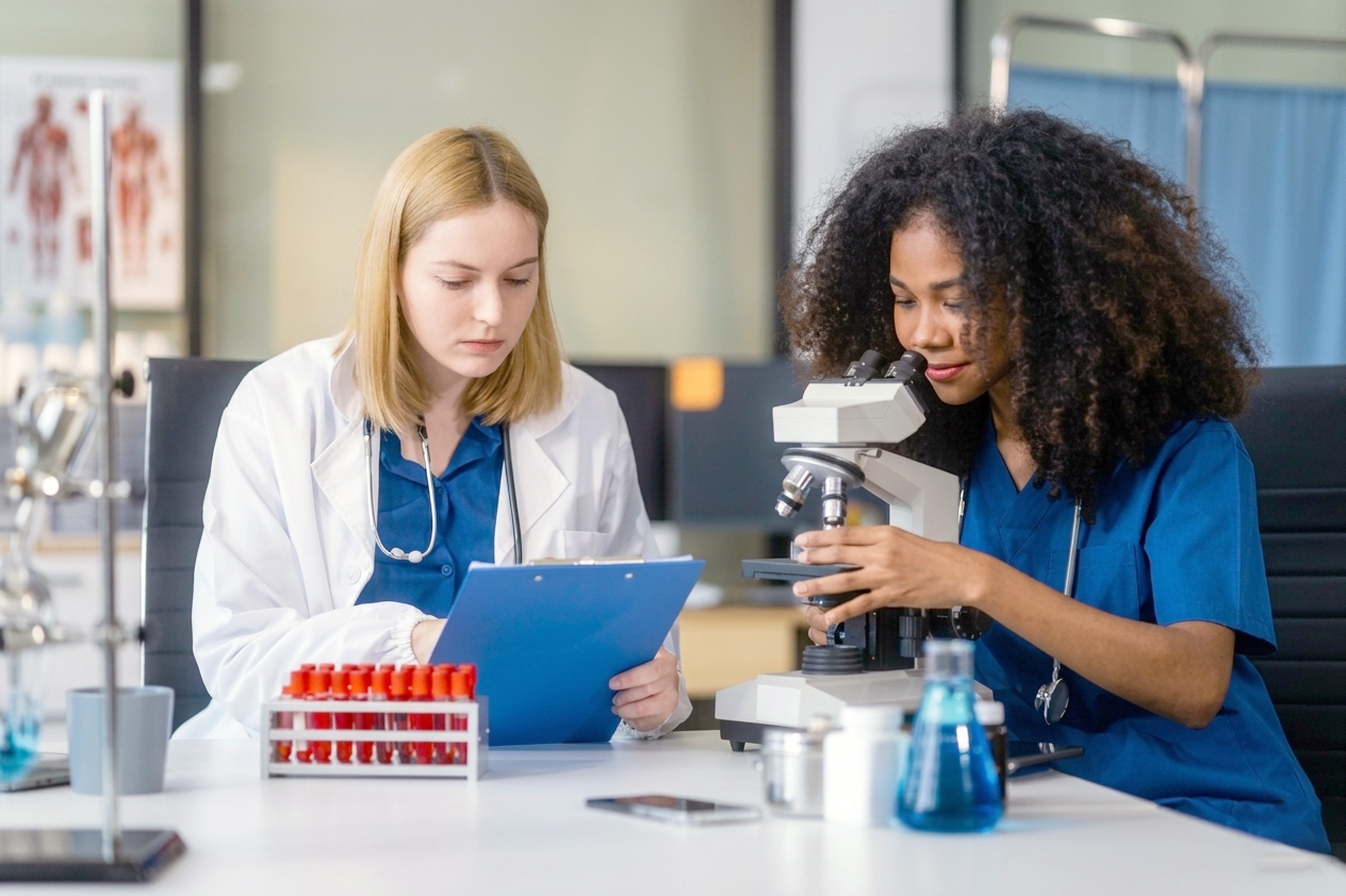A healthcare professional in a white lab coat reviews a clipboard while a colleague in blue scrubs looks through a microscope in a medical laboratory, with blood sample tubes visible on the table.
