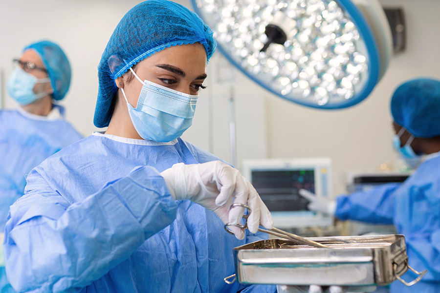 A sterile processing or surgical technician in blue scrubs, a surgical gown, mask, hair net, and gloves handles a tray of surgical instruments in an operating room, with other masked medical personnel and equipment visible in the background.