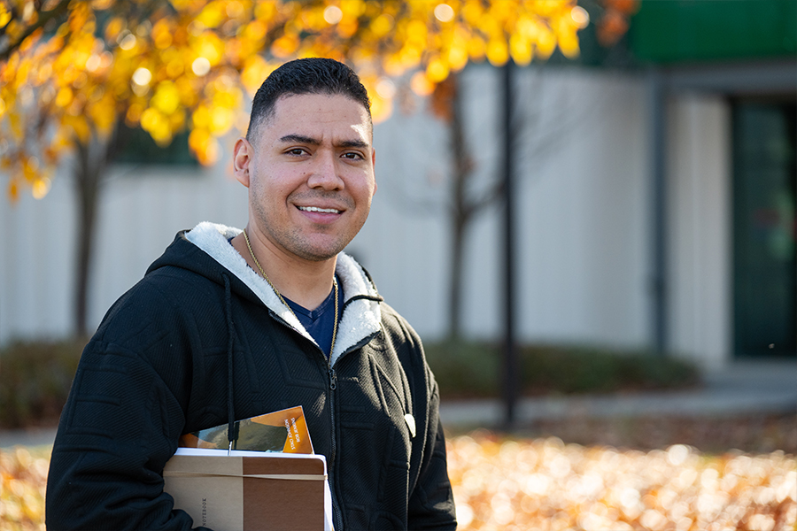 A medium shot of a smiling man outdoors during autumn. He has short dark hair and is wearing a black zip-up hoodie with a white sherpa-lined hood over a blue shirt. He is holding a notebook and papers under his arm. The background is softly blurred, showing a building and trees with golden yellow autumn leaves.