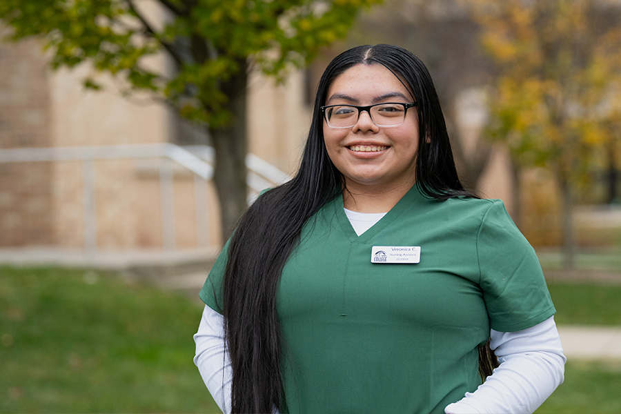 A smiling person with long hair and glasses in a green uniform stands outdoors, with blurred autumn trees and a building in the background.
