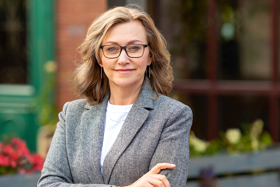 A woman with blonde hair and glasses wearing a gray blazer stands with her arms crossed, smiling confidently outdoors in front of a blurred storefront with flowers and greenery.