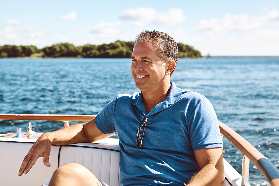 A smiling man in a blue polo shirt relaxes on a boat, with a wooded shoreline and open water in the background.
