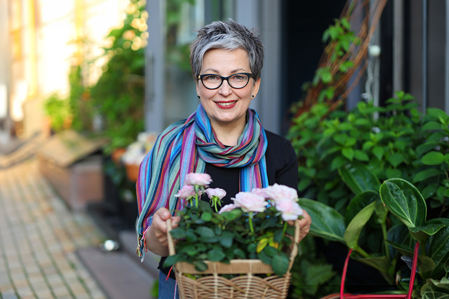 A smiling woman with short gray hair and glasses, wearing a colorful striped scarf, holds a basket of pink roses at an outdoor flower shop