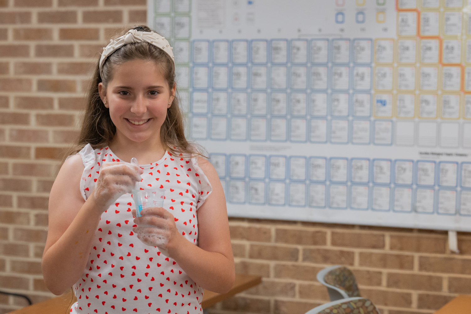 Young girl smiling and holding a foaming science experiment in front of a periodic table.