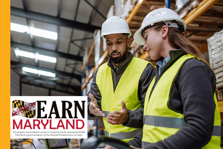 Two warehouse workers in hard hats and safety vests reviewing equipment together, with EARN Maryland grant program logo overlaid in the corner.