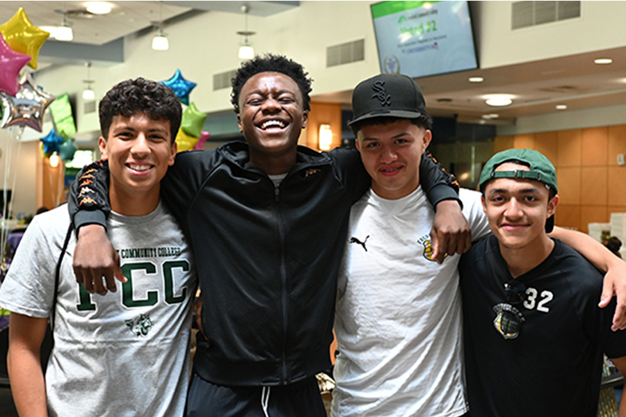 Four smiling young men stand with their arms around each other at an indoor campus event. They are dressed casually, with one wearing a grey Frederick Community College t-shirt and another a green baseball cap, while colorful star-shaped balloons are visible in the background.