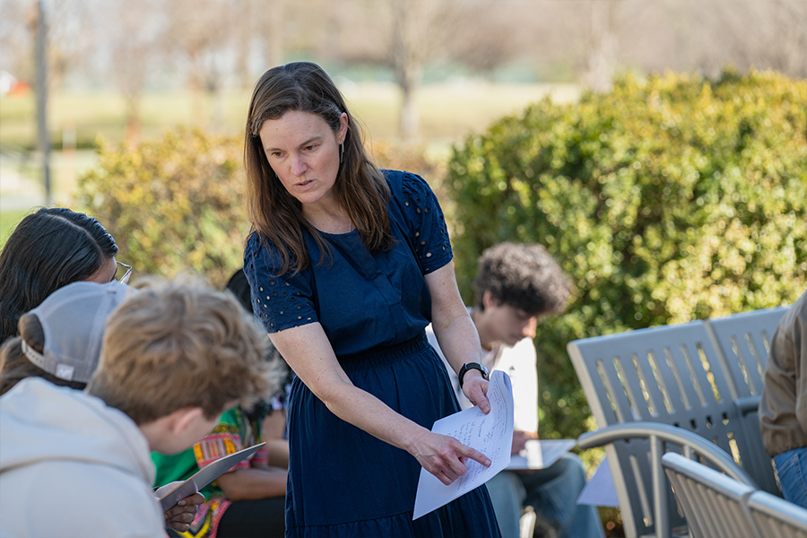 A faculty member in a navy blue dress stands outdoors, pointing to a document and speaking with a group of students seated on a campus patio.