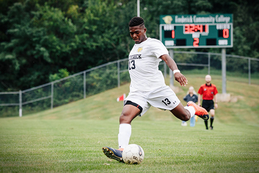 A Frederick Community College men’s soccer player in a white jersey with the number 13 is captured in mid-kick on a grassy field. In the background, a digital scoreboard shows a time of 32:41, with FCC leading 1-0. A referee in a red shirt stands nearby, and the field is surrounded by lush green trees and a chain-link fence.