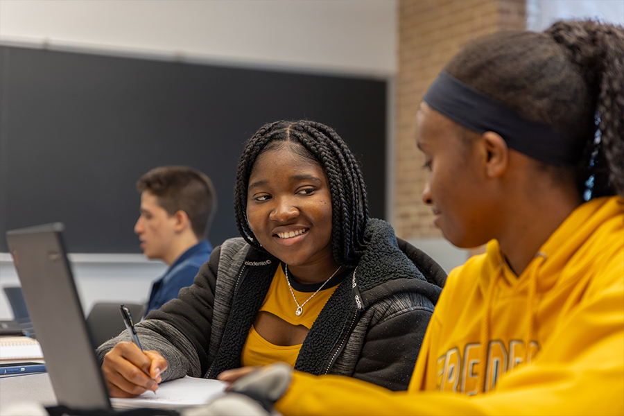 Two students collaborate at a table in a classroom. One student with braided hair smiles while writing in a notebook and looking at a laptop screen.