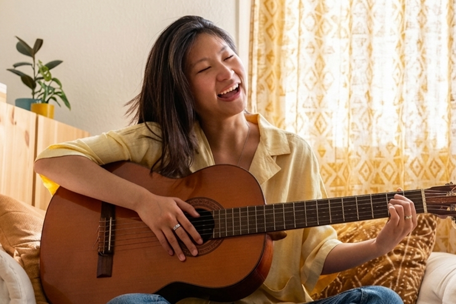 A young woman in a yellow shirt laughs joyfully while playing an acoustic guitar on a couch in a bright, warmly lit living room.