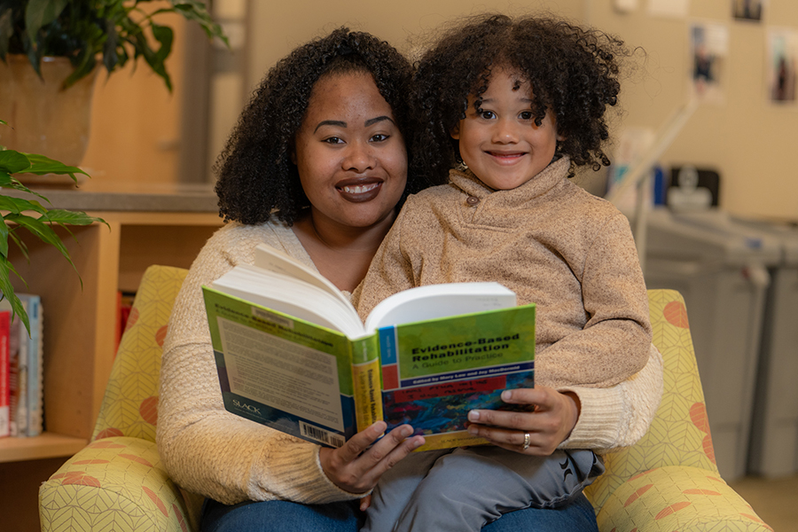 A smiling student-parent sits in a chair indoors, holding a young child in her lap while they look at an open textbook together.