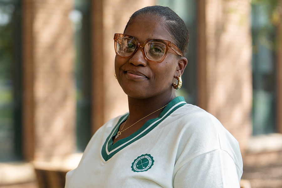 A portrait of a smiling woman with glasses and gold hoop earrings standing outdoors in front of a brick campus building.