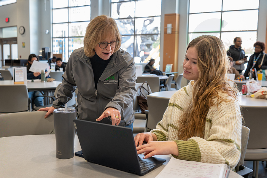 A staff member in a grey college-branded jacket points to a laptop screen while assisting a student seated at a round table in a bright, modern campus common area. Both are focused on the screen in a collaborative and supportive manner.