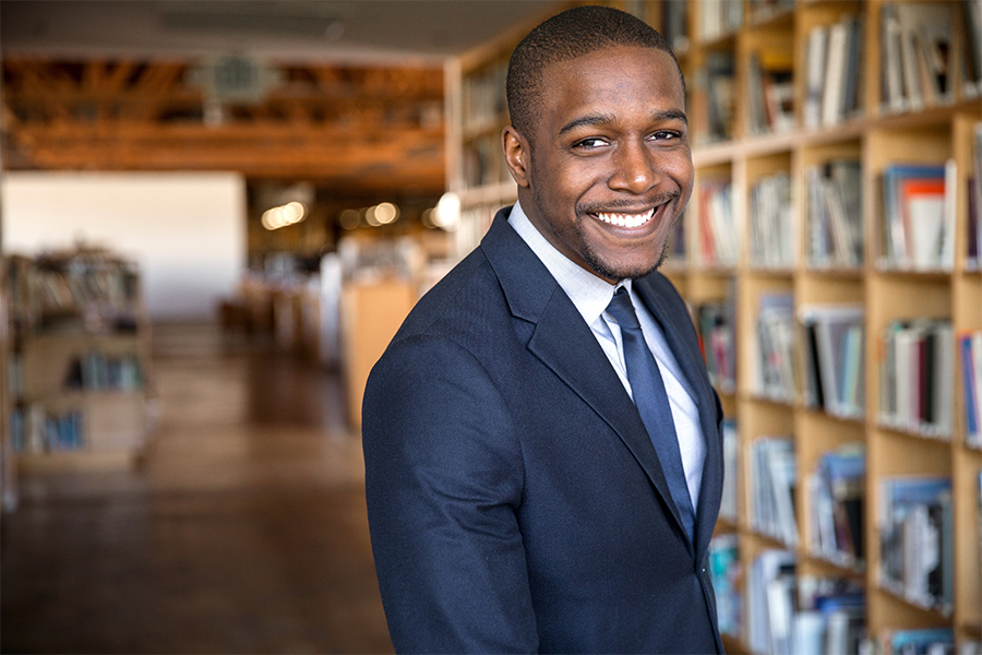 A portrait of a smiling young Black man in a dark suit, light blue shirt, and tie. He is standing in a brightly lit library with floor-to-ceiling wooden bookshelves filled with books in the background.