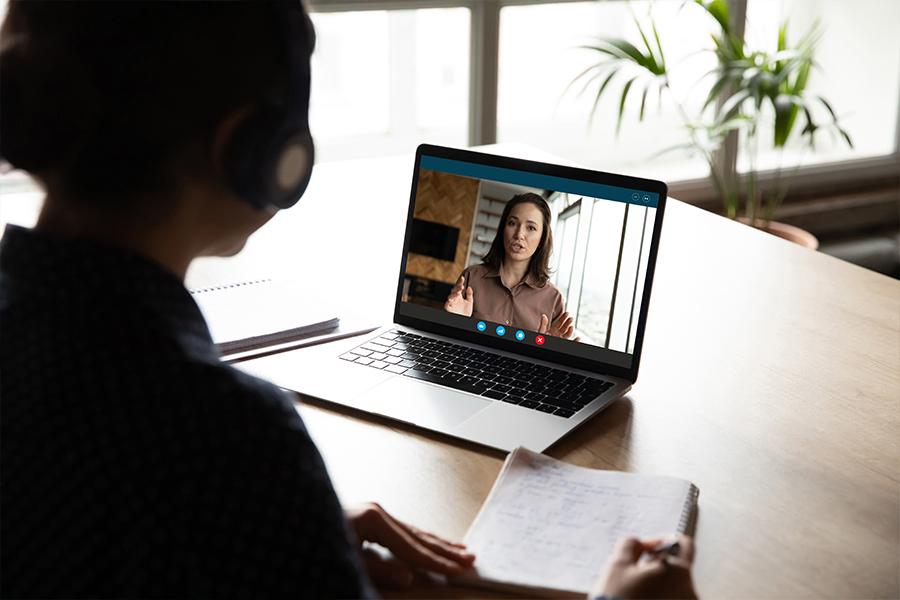 An individual with dark hair tied back wears large black headphones and sits at a desk, viewed from behind. They are facing a laptop that shows a woman on a video call. The woman on the screen has brown hair and is gesturing with her hands while speaking.