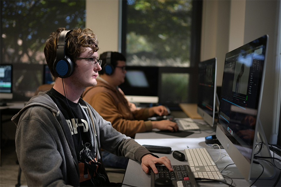 Two students with headphones work at a row of computers in a bright classroom, focused on editing video content on large monitors.