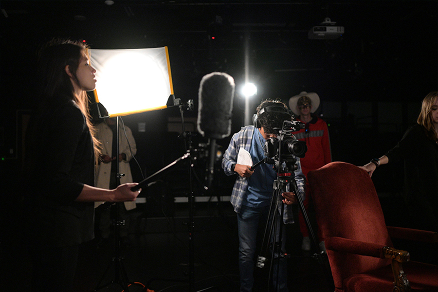 Behind-the-scenes view of a film set where students operate a camera on a tripod, a boom mic, and a large studio light in a dark room.