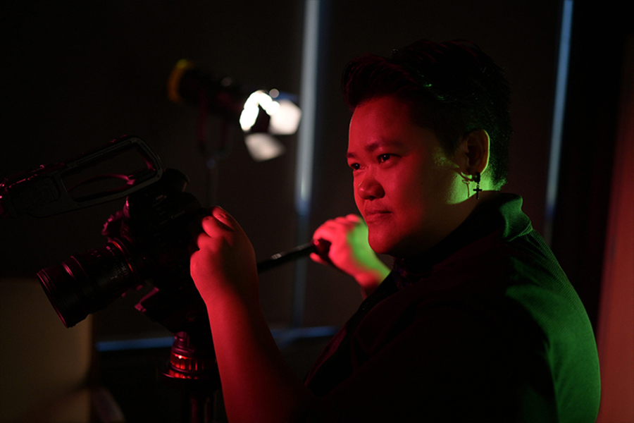 Close-up of a student filmmaker operating a camera, their face dramatically lit by vibrant red and green production lights in a dark studio.