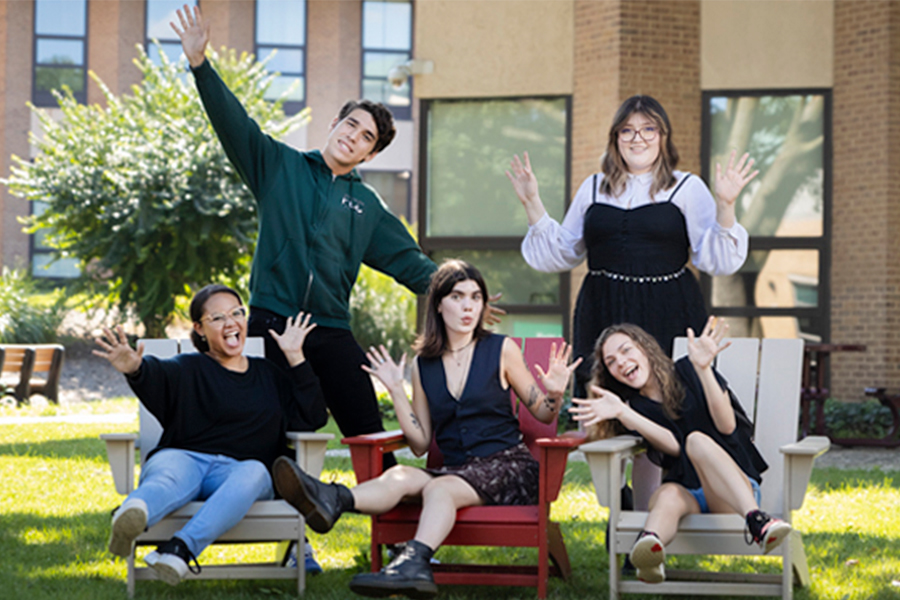 Five students pose playfully on a grassy lawn in front of a brick campus building. Three students are seated in Adirondack chairs—two in grey chairs flanking one in a red chair—while two other students stand behind them. All five are waving their hands and making enthusiastic expressions toward the camera. One student wears a green hoodie featuring the FCC logo.