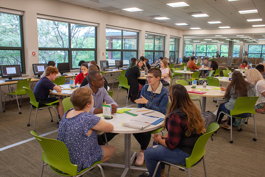 A high-energy collaborative study space with several groups of students and instructors seated at round white tables with bright green chairs. Large windows line the left wall, and the background shows a row of computer workstations. The atmosphere is academic and interactive.