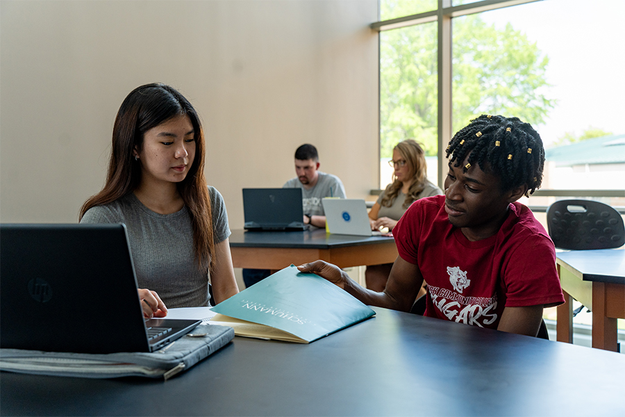 A group of diverse college students studying together at a desk. In the foreground, a female student looks at a laptop while a male student in a Cougars t-shirt points to a teal folder. Other students work on laptops in the background in a bright, sunlit room.