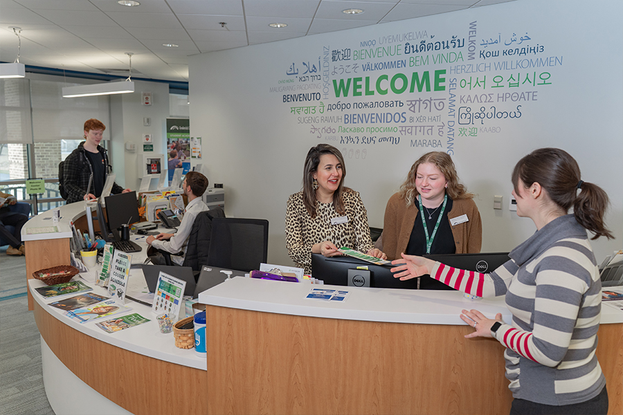 Two staff members stand behind a curved wooden welcome desk, assisting a student. One staff member in a leopard print blouse hands a document to the student, while the other in a brown cardigan looks on. In the background, a large wall graphic displays the word "WELCOME" in many different languages and colors. Other staff members and students are visible further down the counter in the bright, modern office space.