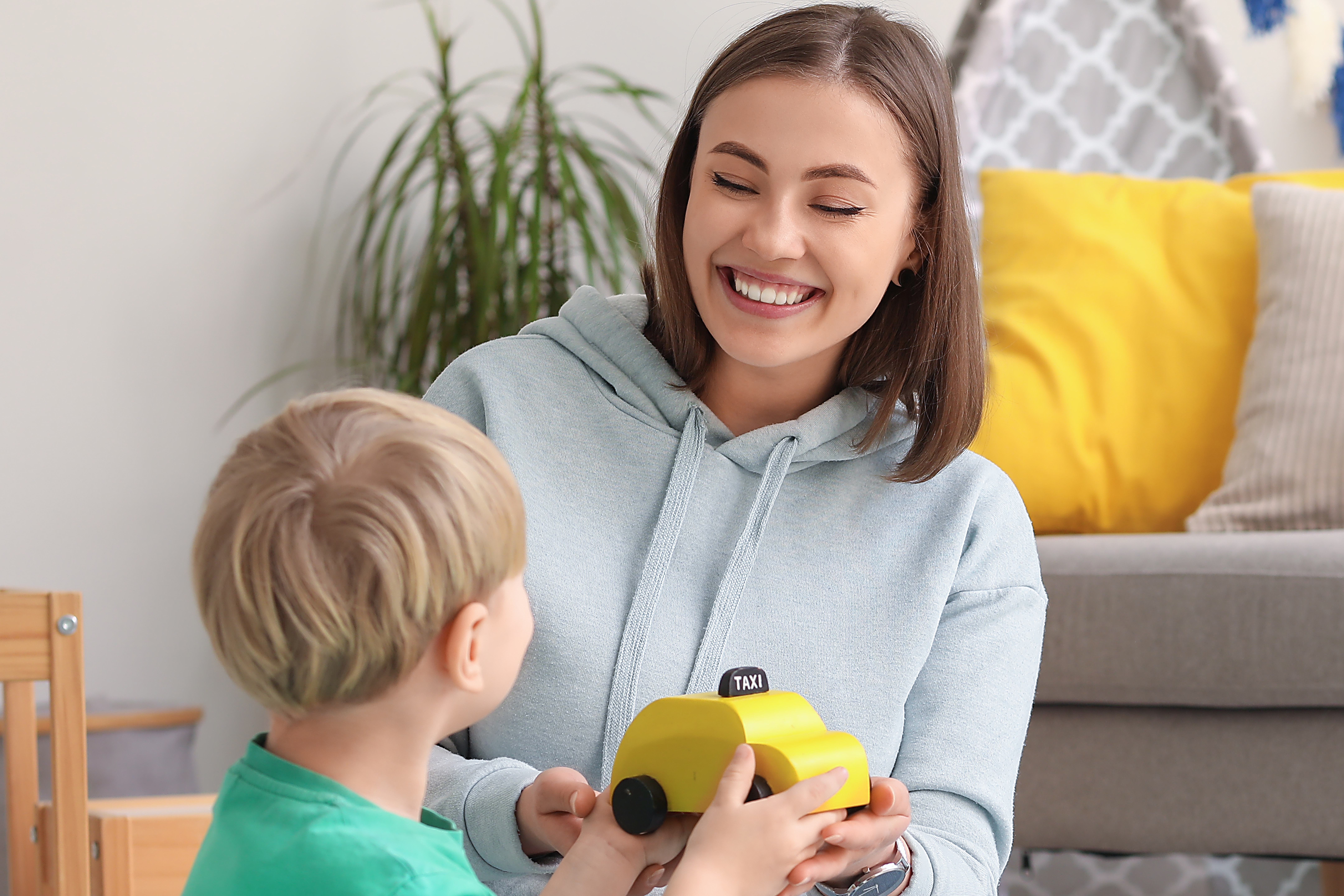 smiling young woman plays with a toddler on the floor of a living room, both holding a yellow wooden toy taxi cab. A couch with a yellow pillow and a houseplant are visible in the background.