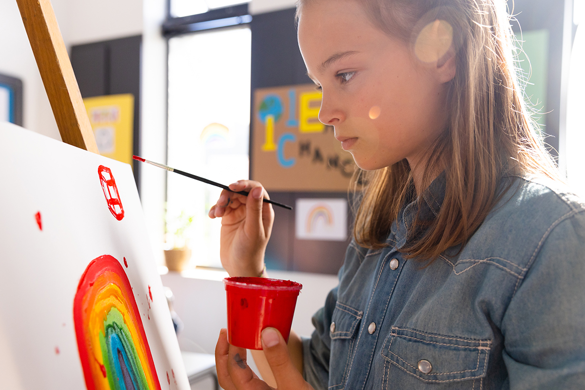 A girl focuses intently on painting a rainbow on a canvas at an easel, holding a small cup of red paint and a fine paintbrush. Colorful artwork is visible on the wall behind her.