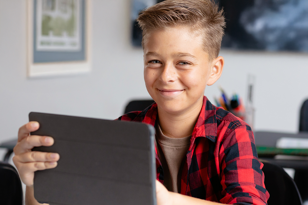 A smiling boy holds up a tablet in a classroom, looking pleased. A cup of pencils and framed artwork are visible in the background.