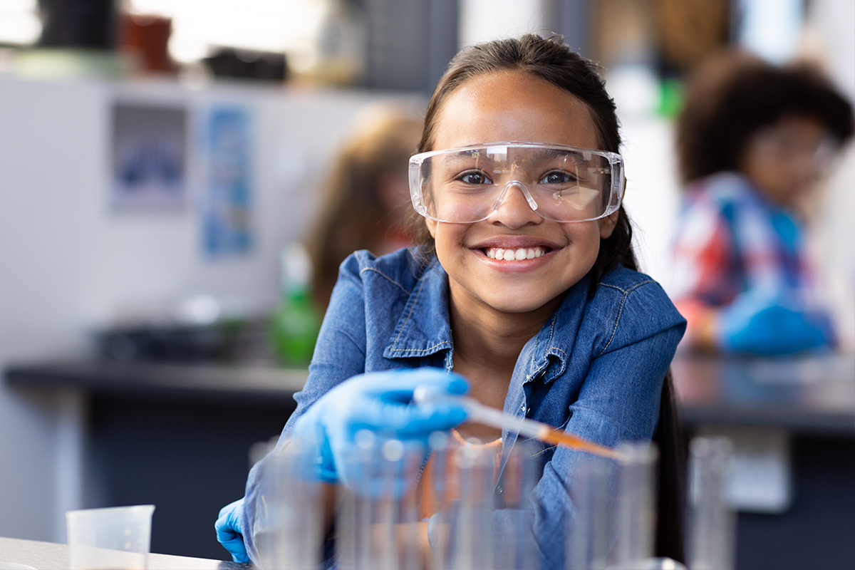 A girl wearing safety goggles and blue lab gloves smiles while using a dropper over a row of test tubes in a science classroom. Another student works in the background.